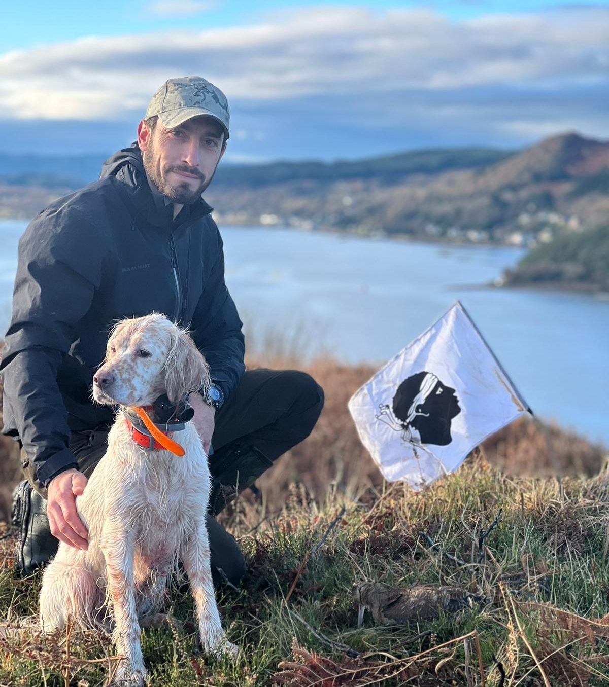Homme avec un chien assis, près d'un drapeau, surplombant un lac et des collines.